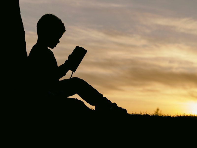 A child reading a book outdoors, in silhouette against an evening sky