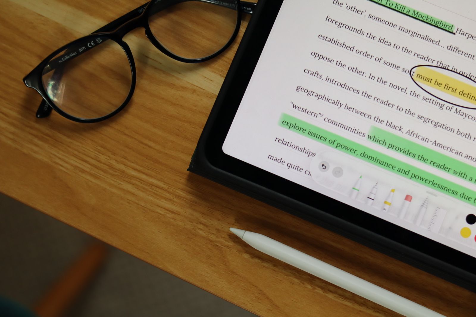 An essay on To Kill a Mockingbird on an iPad during marking, with a phrase circled and lines highlighted, beside an Apple Pencil and a pair of glasses.
